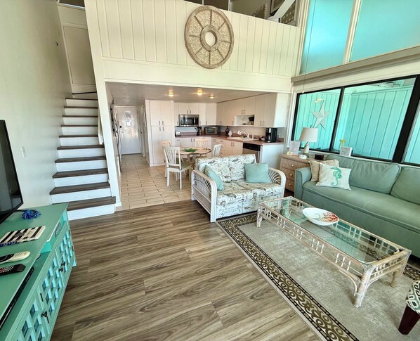 A spacious living area with a high ceiling, a staircase, a kitchen, and a dining area.