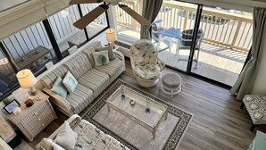 A spacious living room with a large window, a ceiling fan, a sofa set, a coffee table, and a patterned rug.