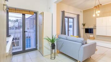 Un appartement moderne avec un canapé gris, une plante en pot et un balcon offrant une vue sur les immebles.