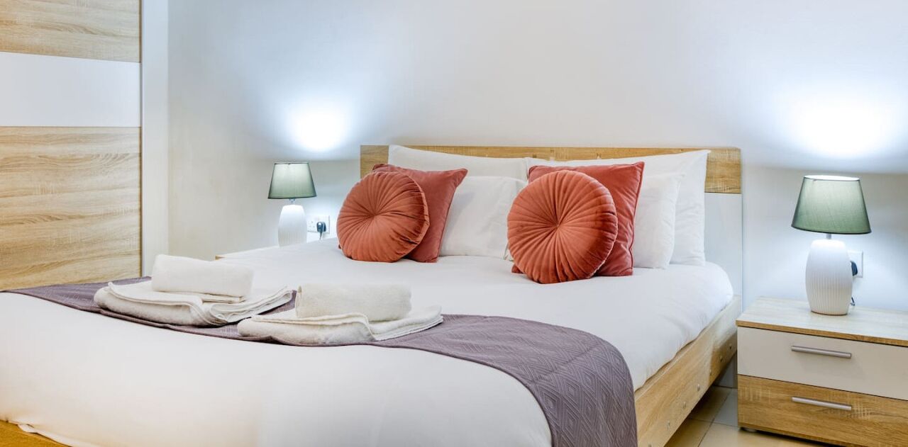 Una cama bien tendida con sábanas blancas y dos almohadas decorativas rosas, un cabecero de madera y dos lámparas de noche.
