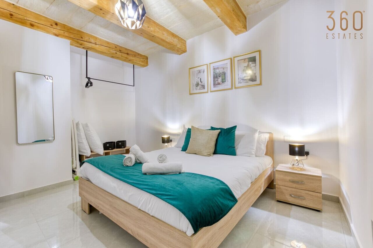 Une chambre moderne avec un lit en bois, des murs blancs et un plafond avec des poutres apparentes. On y trouve trois tableaux encadrés au mur, une table de chevet avec une lampe et un miroir.
