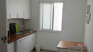 Una cuina compacta amb armaris blancs, un rentaplats integrat, una finestra amb persianes i una taula de dinar petita amb cadires.
