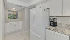 Una cocina con armarios blancos, un refrigerador, una cafetera y una ventana con vista a follaje.