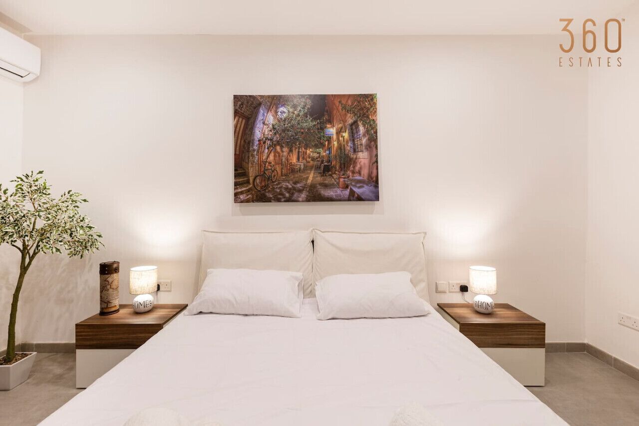 A bedroom with a large bed, two bedside tables with lamps, a potted plant, and a wall-mounted painting.
