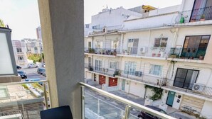 A balcony with two chairs and a table, overlooking a street with parked cars and buildings.