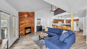 A modern living room with a blue sectional sofa, a wooden accent wall, a fireplace, and a kitchen in the background.