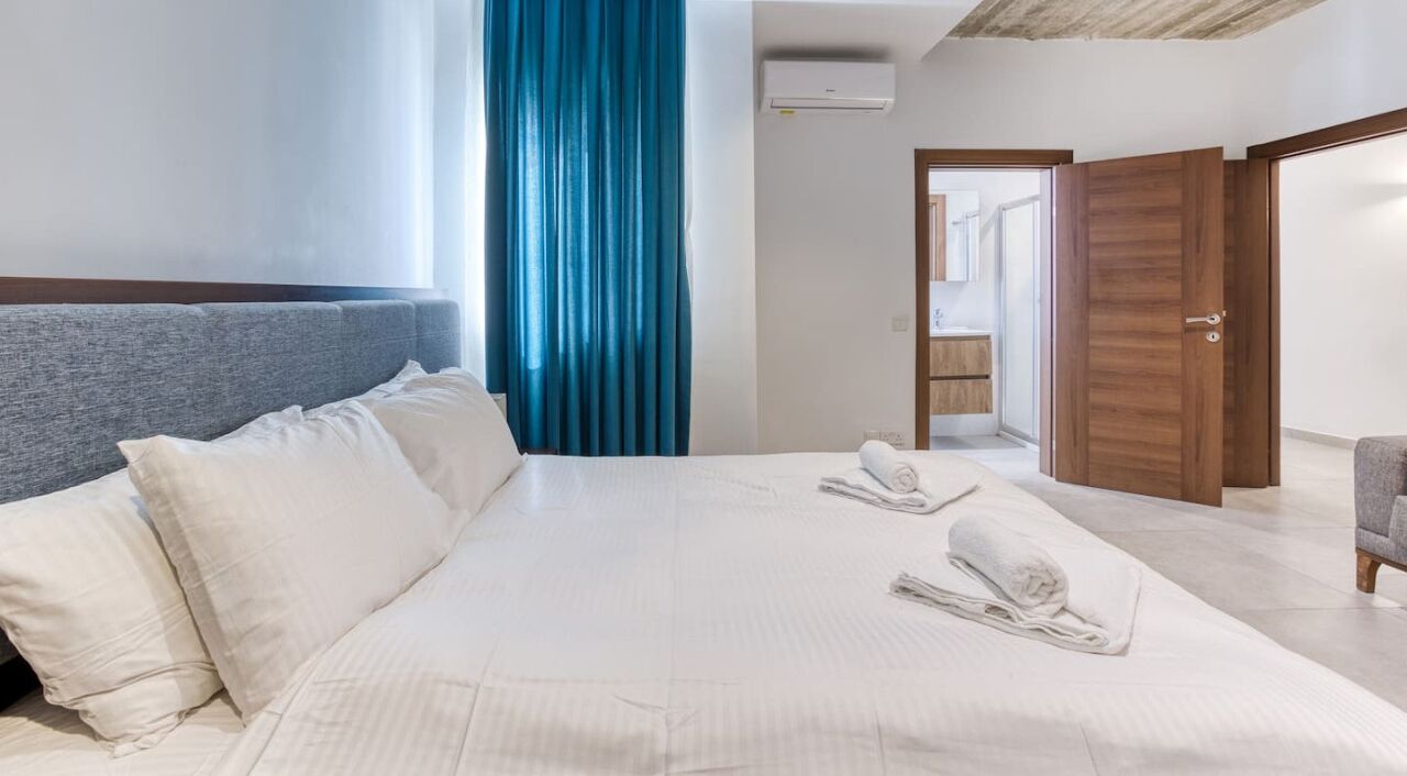Habitación de hotel con una cama grande, cabecera gris, cortina azul y puerta de madera.