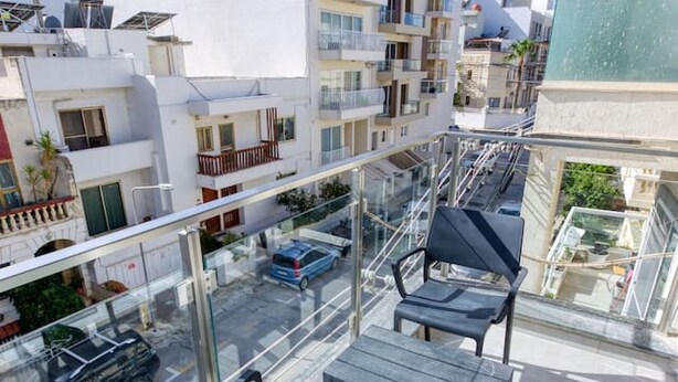 En balkon med moderne møbler, frit udsyn til gaden nedenfor, og en bygning med både altaner og solpaneler.