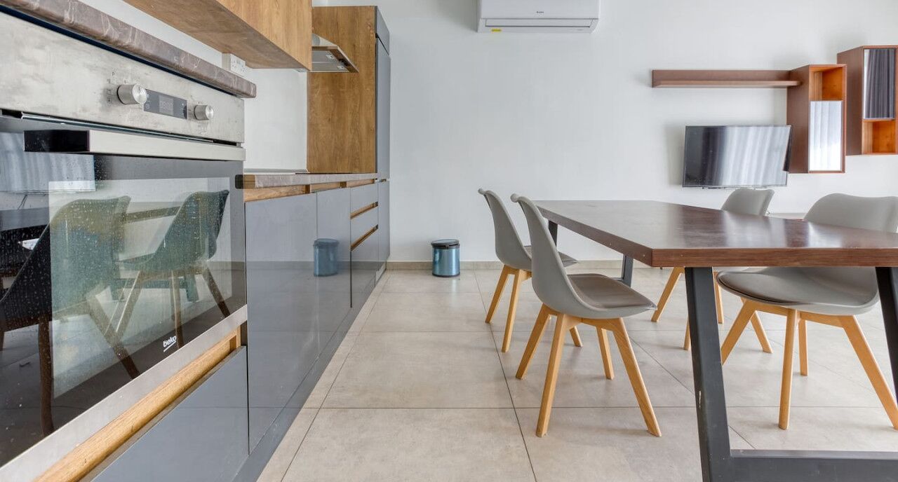 A modern kitchen with a built-in oven, a dining area with a wooden table and chairs, and a TV mounted on the wall.