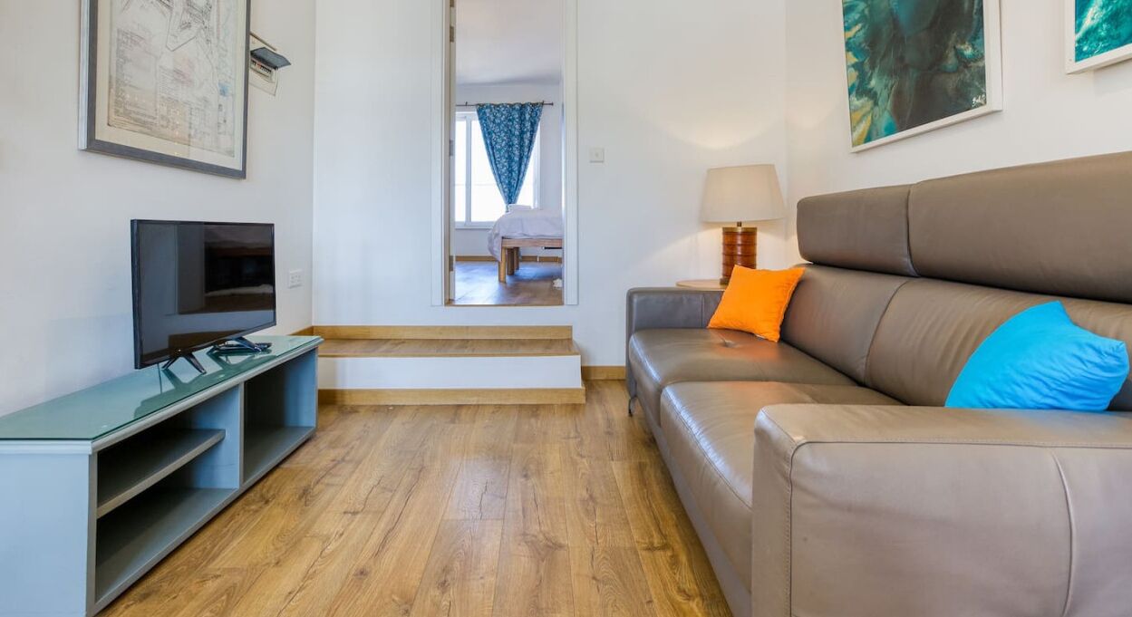 Una sala de estar moderna con un televisor de pantalla plana, un sofá con cojines azules y naranjas, piso de madera y una escalera que conduce a otra habitación.