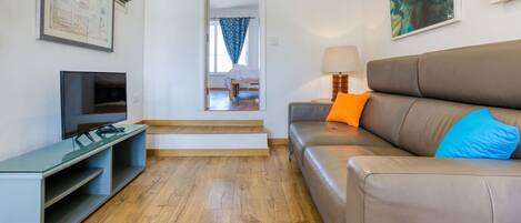 Una sala de estar moderna con un televisor de pantalla plana, un sofá con cojines azules y naranjas, piso de madera y una escalera que conduce a otra habitación.