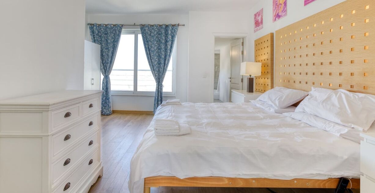 Une chambre à coucher avec une tête de lit en bois, un lit blanc, une table de chevet, une commode et une fenêtre avec des rideaux.