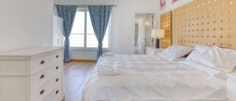Une chambre à coucher avec une tête de lit en bois, un lit blanc, une table de chevet, une commode et une fenêtre avec des rideaux.