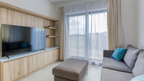 Un salon moderne doté d’un téléviseur à écran plat, de meubles en bois, d’un canapé gris agrémenté de coussins bleus et d’une grande fenêtre ornée de rideaux légers.