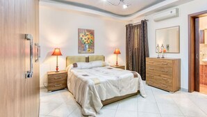 A bedroom with a bed, two bedside tables with lamps, a dresser, a painting on the wall, and a ceiling light fixture.
