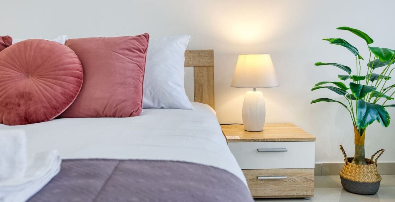 A neatly made bed with a wooden headboard, a bedside lamp, a small drawer, and a potted plant on a bedside table.