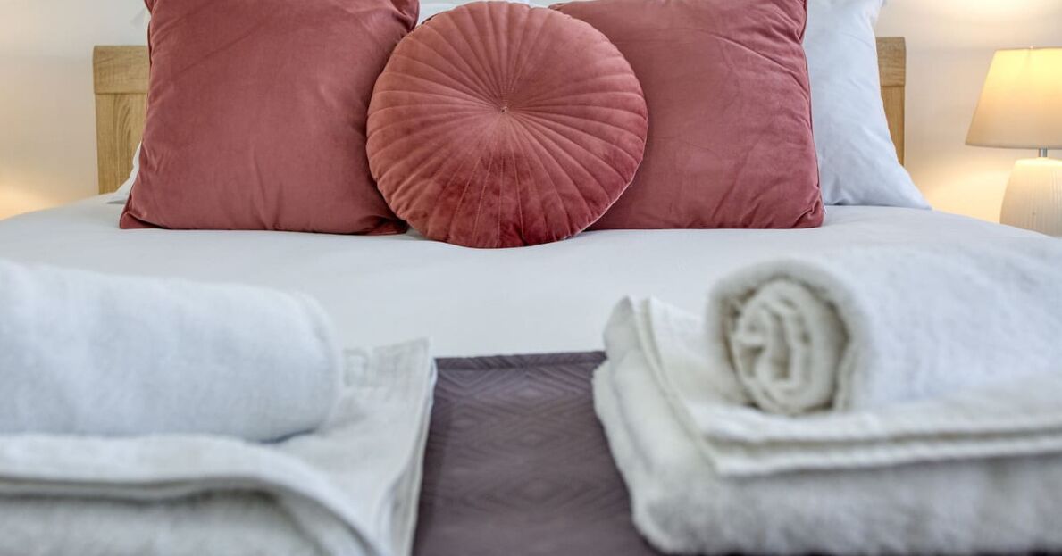 A neatly made bed with pink and white pillows, a folded white towel, and a rolled white towel on a bed with a wooden headboard.