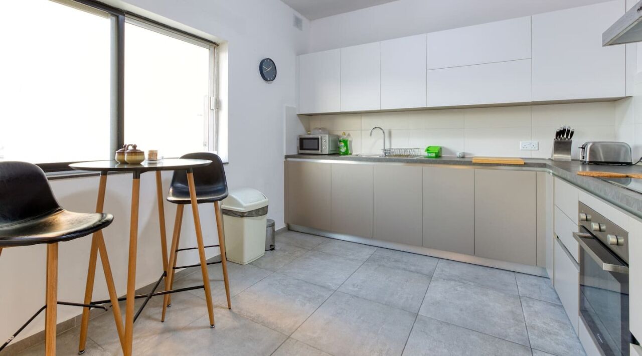 A modern kitchen with white cabinets, a microwave, a sink, and a toaster. There are two black chairs and a small table.