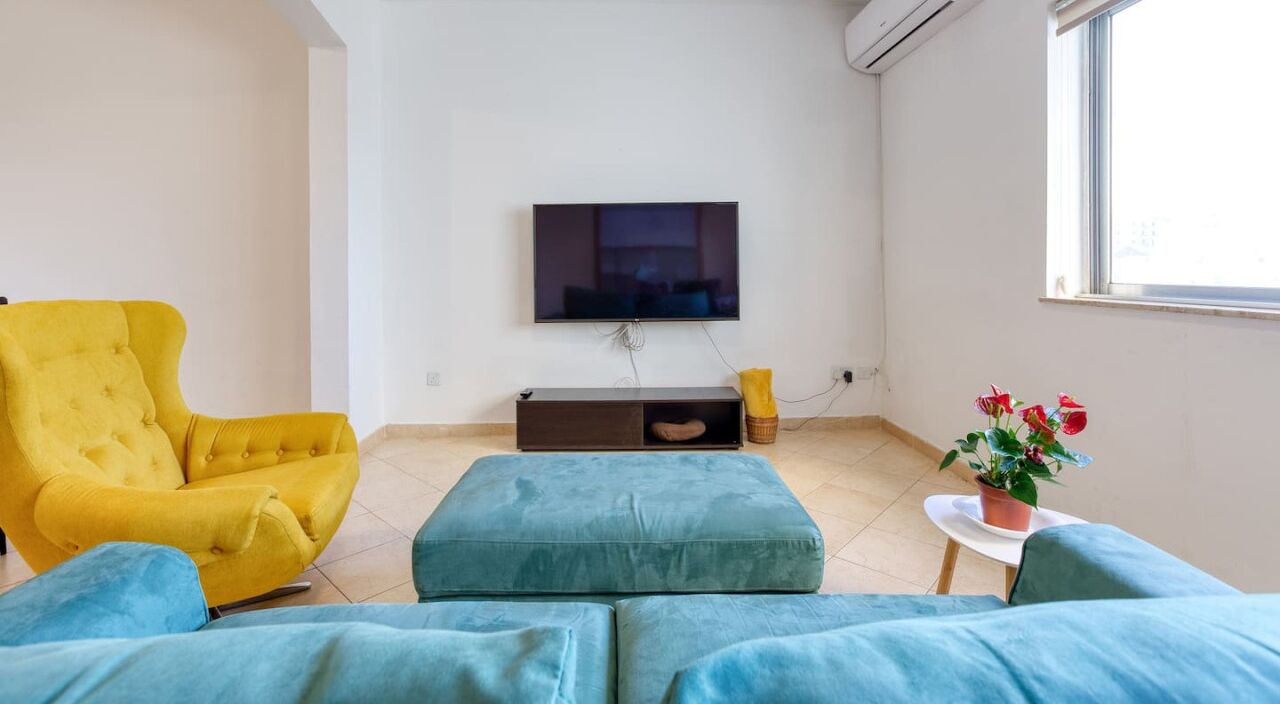 Una sala de estar con un sillón amarillo, un sofá azul, un televisor de pantalla plana montado en la pared y una mesita con una planta en maceta.