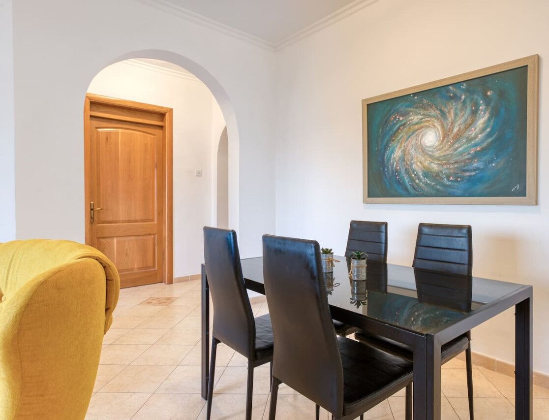 A dining area with a table and chairs, a painting on the wall, and a wooden door in the background.