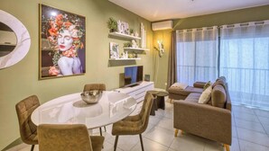 A modern living room with a dining area, featuring a sofa set, a round dining table, and a large floral artwork on the wall. - Modern & private 1BR APT in the Heart of Paceville (St. Julian`s)