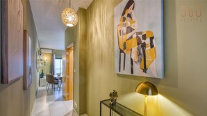 A hallway with a modern design, featuring a glass console table with a lamp, a large abstract painting, and a dining area in the background. - Modern & private 1BR APT in the Heart of Paceville (St. Julian`s)