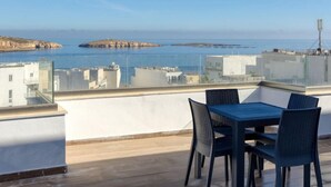 Une terrasse sur le toit, avec une table et des chaises, offrant une vue sur un paysage côtier avec des bâtiments et des falaises.