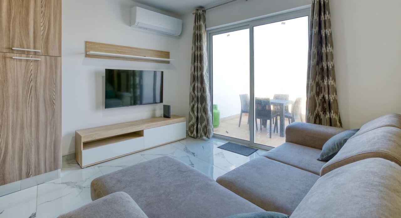 Een moderne woonkamer met een flatscreen televisie, grijze banken, een houten kast en een glazen schuifdeur naar een buitenruimte.