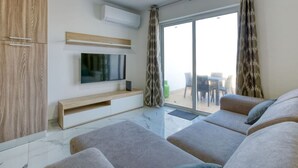 A modern living room with a flat-screen TV, grey sofas, a wooden cabinet, and a sliding glass door leading to an outdoor area.