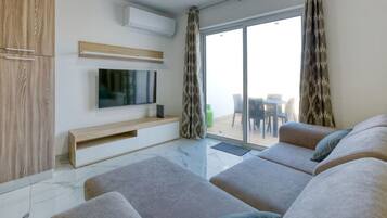 Een moderne woonkamer met een flatscreen televisie, grijze banken, een houten kast en een glazen schuifdeur naar een buitenruimte.