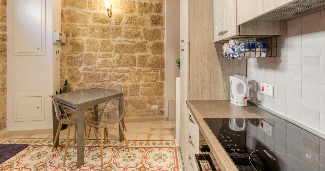 A kitchen with a stone wall, a table with chairs, a kettle, and a built-in oven.