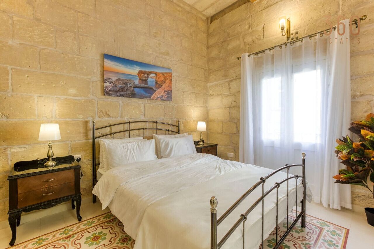A bedroom with a stone wall, a bed with white linens, two bedside tables with lamps, and a painting of a bridge.