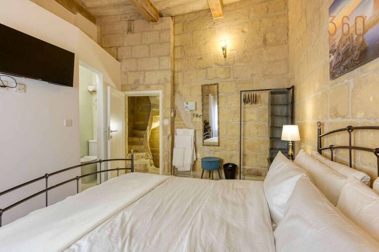 Una camera da letto con parete in pietra, un letto con lenzuola bianche, un comodino con una lampada e uno specchio appeso al muro.