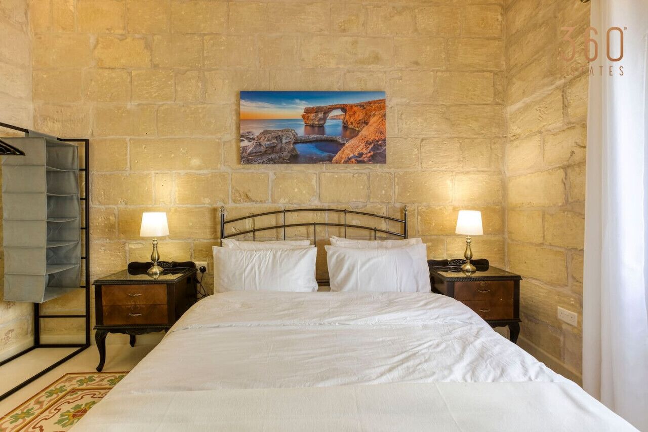 A hotel room with a stone wall, a bed with white linens, two bedside tables with lamps, and a painting of a coastal scene.