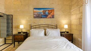 A hotel room with a stone wall, a bed with white linens, two bedside tables with lamps, and a painting of a coastal scene.