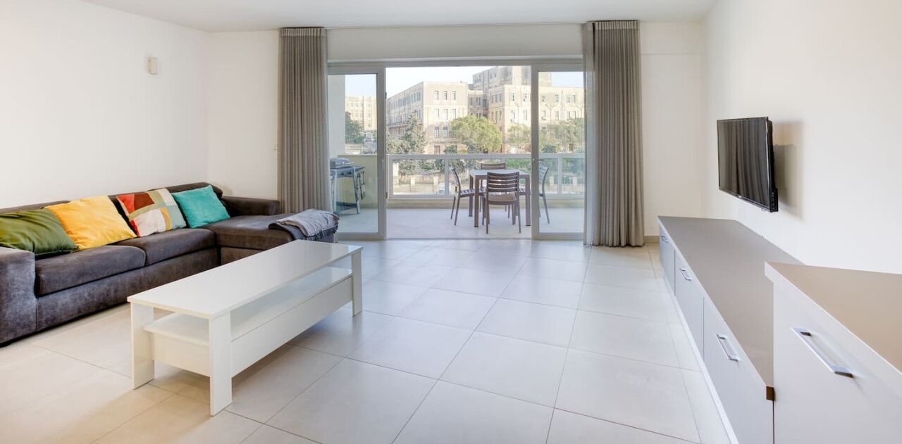A modern living room with a sofa, a white coffee table, a TV mounted on the wall, and a balcony with a table and chairs.