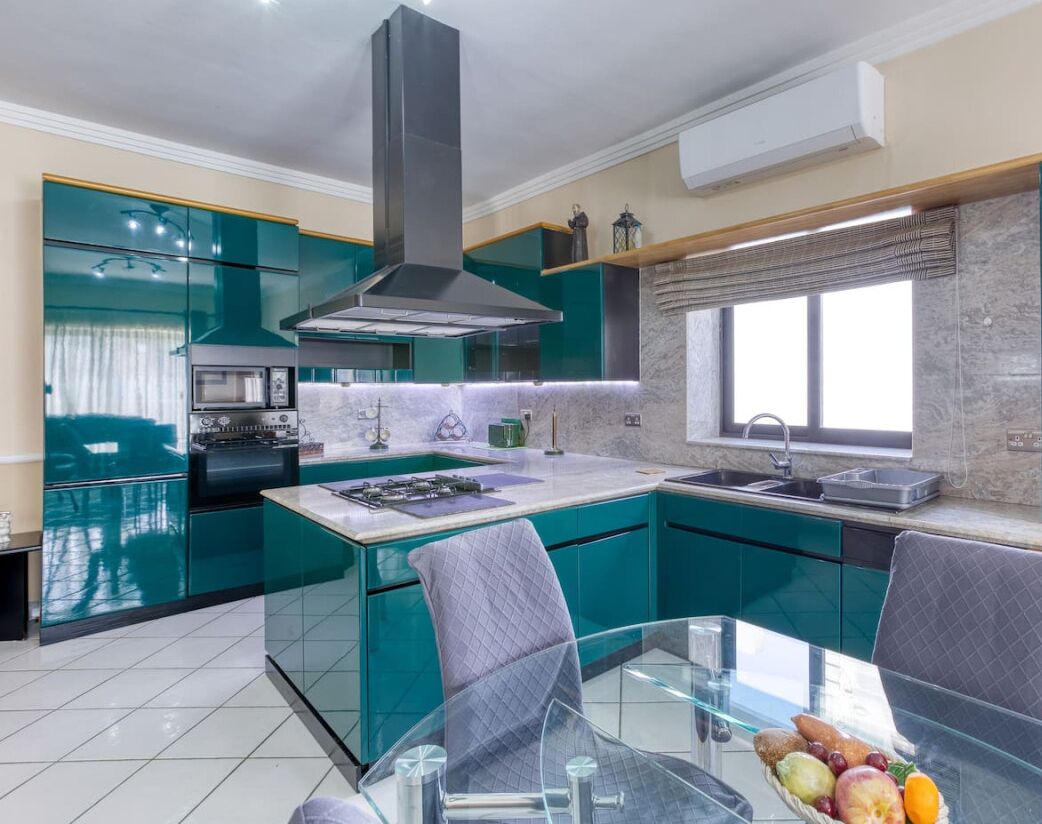 A modern kitchen with teal cabinetry, a central island, and a dining area with a glass table and chairs.