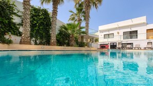 Une piscine à l’eau d’un bleu cristallin, entourée de palmiers et bordée d’un bâtiment moderne à la façade blanche.