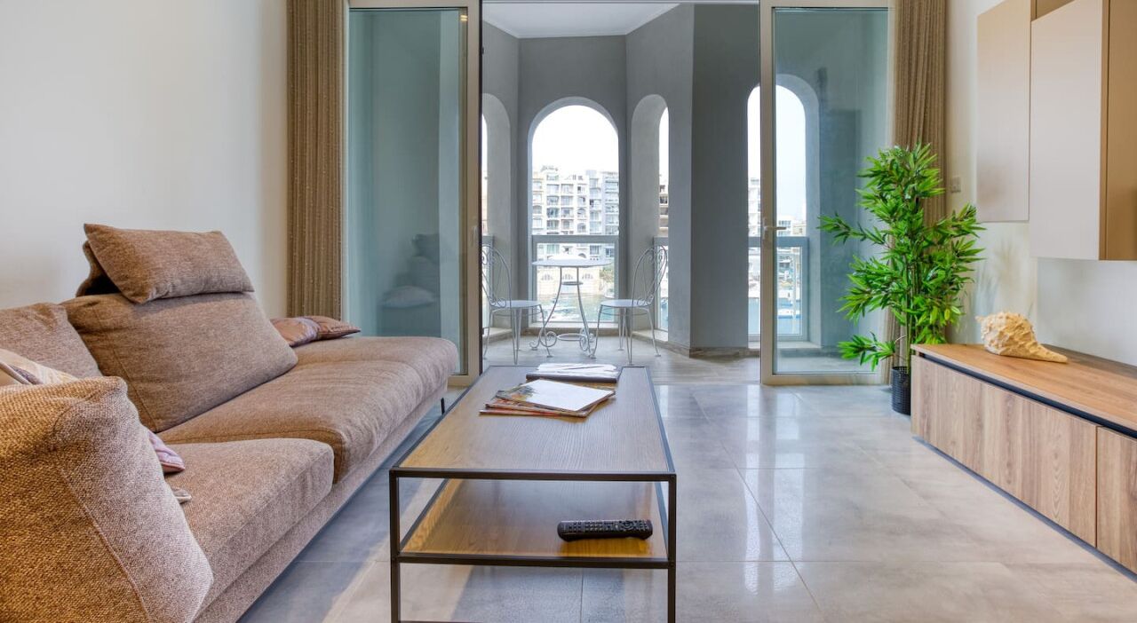A modern living room with a sofa, a coffee table, and a view of a balcony with a pool.