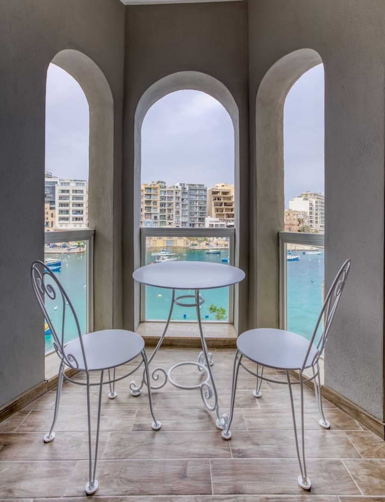 A balcony with two metal chairs and a small round table, overlooking a marina and buildings.