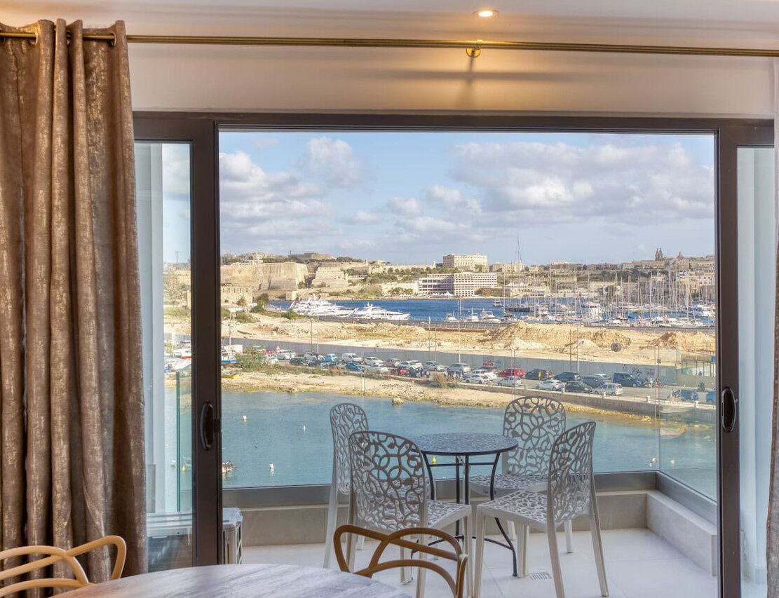 Een balkon met een tafel en stoelen, met uitzicht op een jachthaven met boten en een verre skyline.