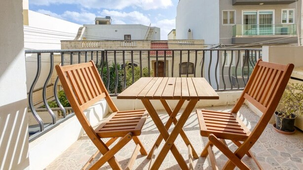 A balcony with a wooden table and two chairs, overlooking a residential area with buildings and a clear sky. - Charming 2BR Apt with Cosy Balcony in Marsascala (Wied il-Għajn)
