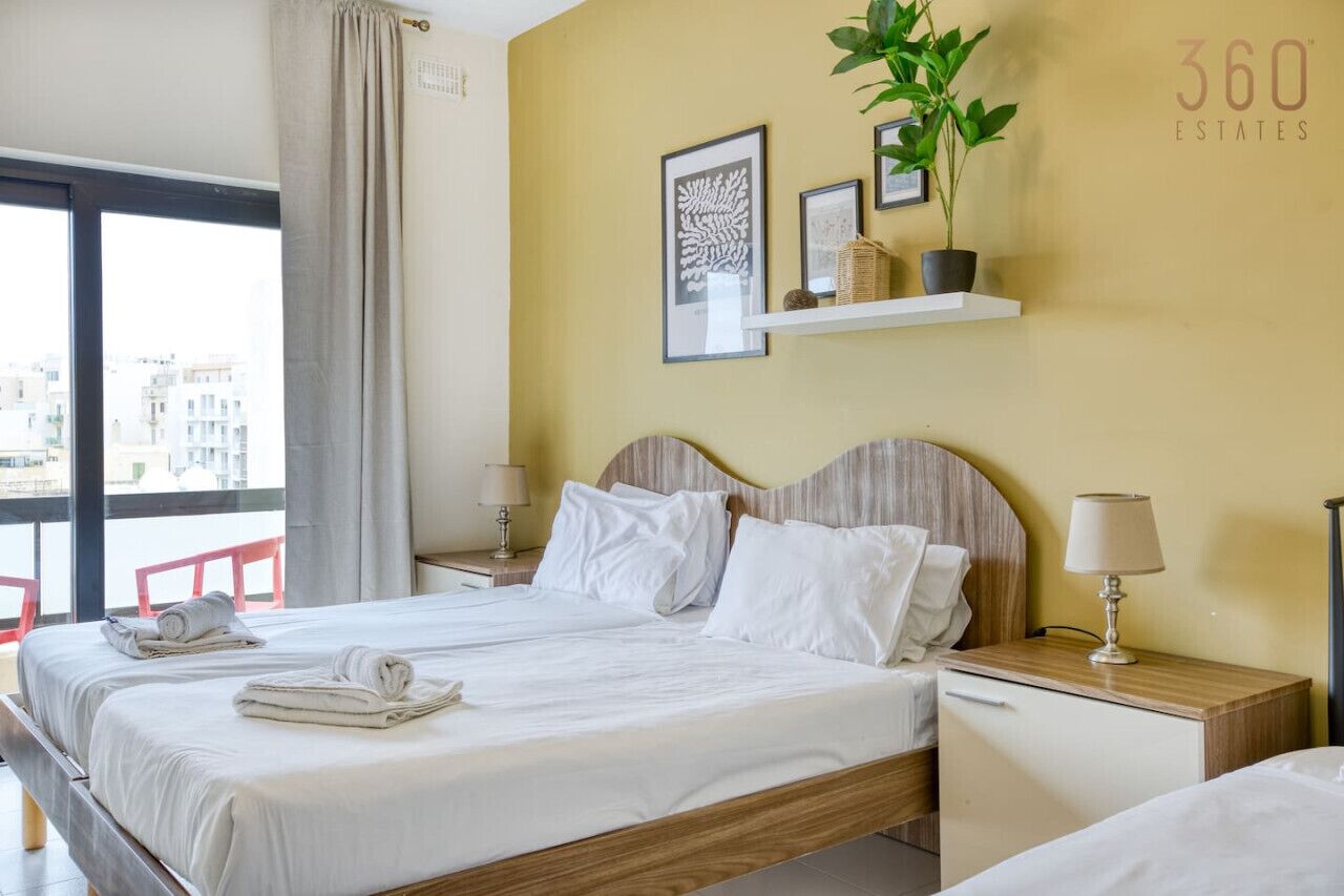 A hotel room with a bed, two bedside tables, a nightstand, a window with a view, and a wall-mounted shelf with decorative items.