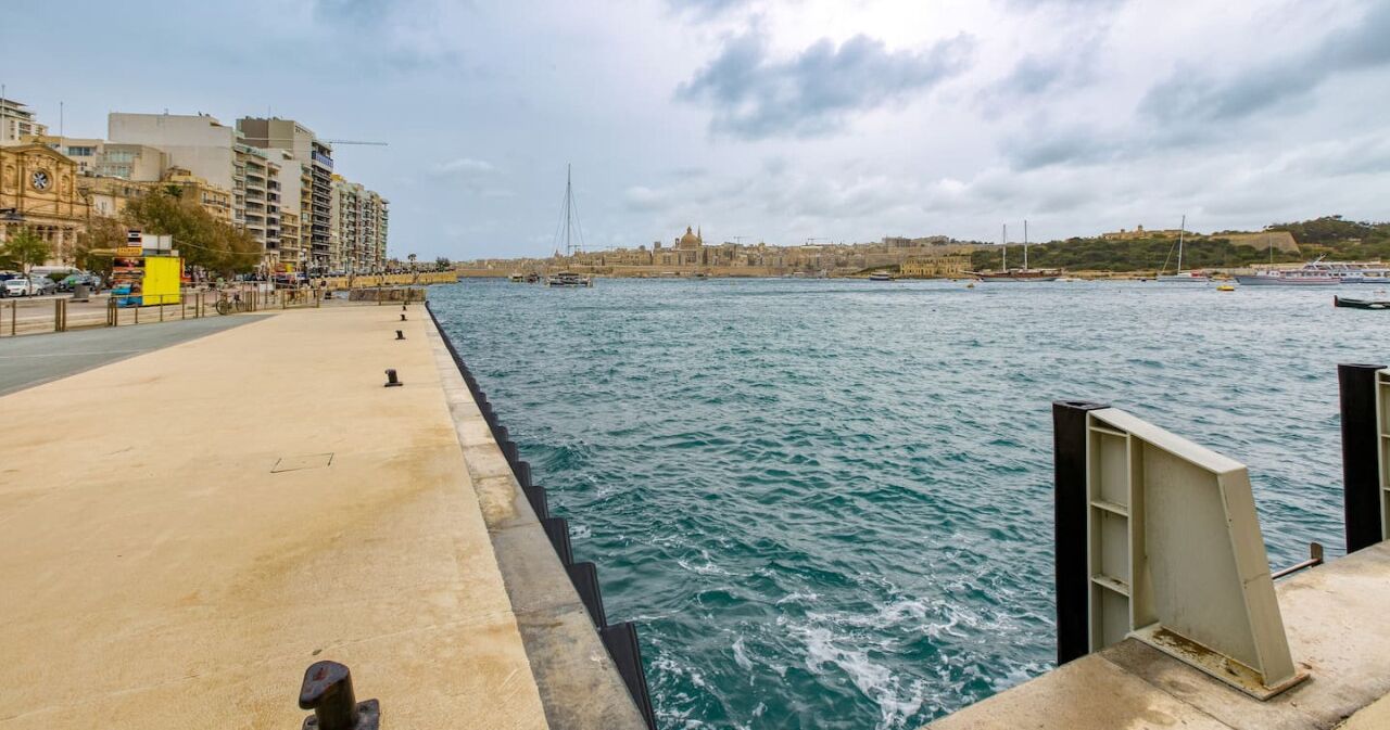 A waterfront promenade with a concrete walkway, a view of the sea, and buildings in the background.