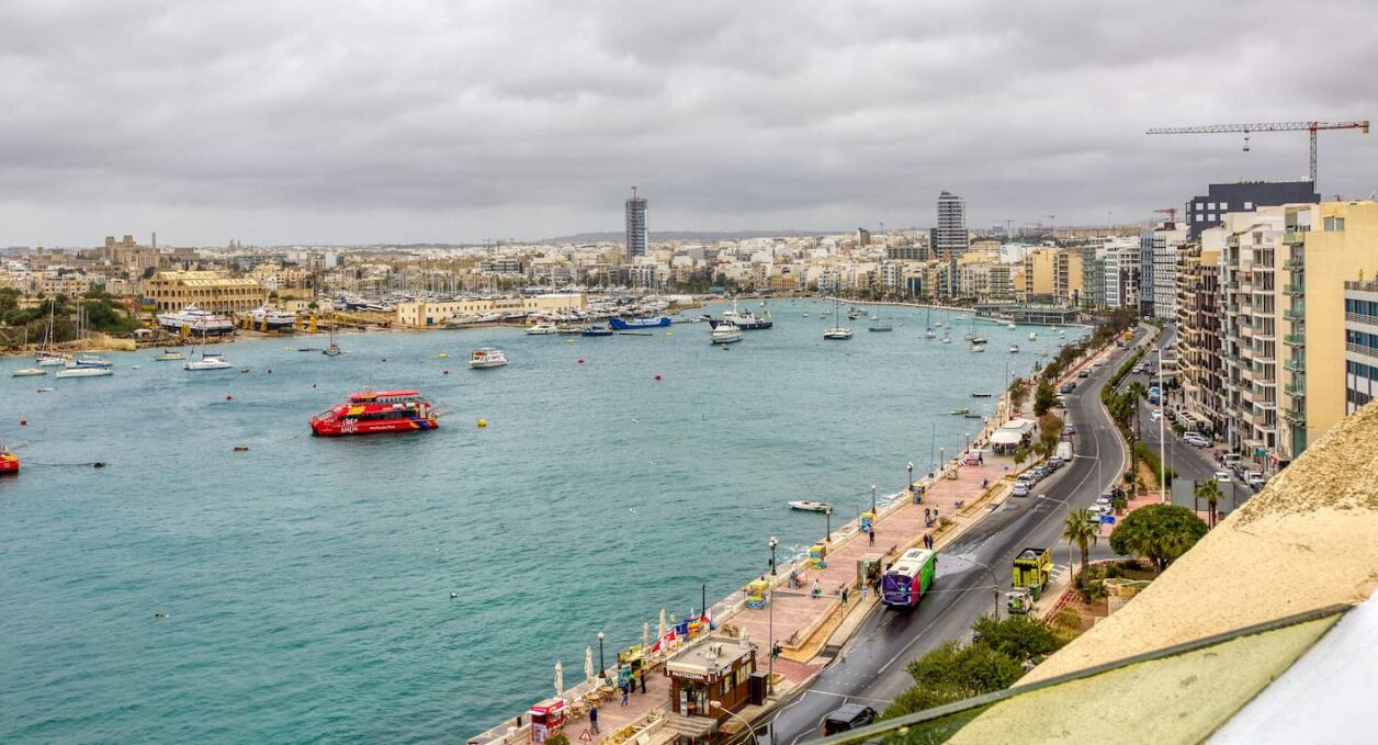 Eine Küstenstadt mit einer Marina voller Boote, einer roten Fähre und einer Fußgängerpromenade am Wasser.