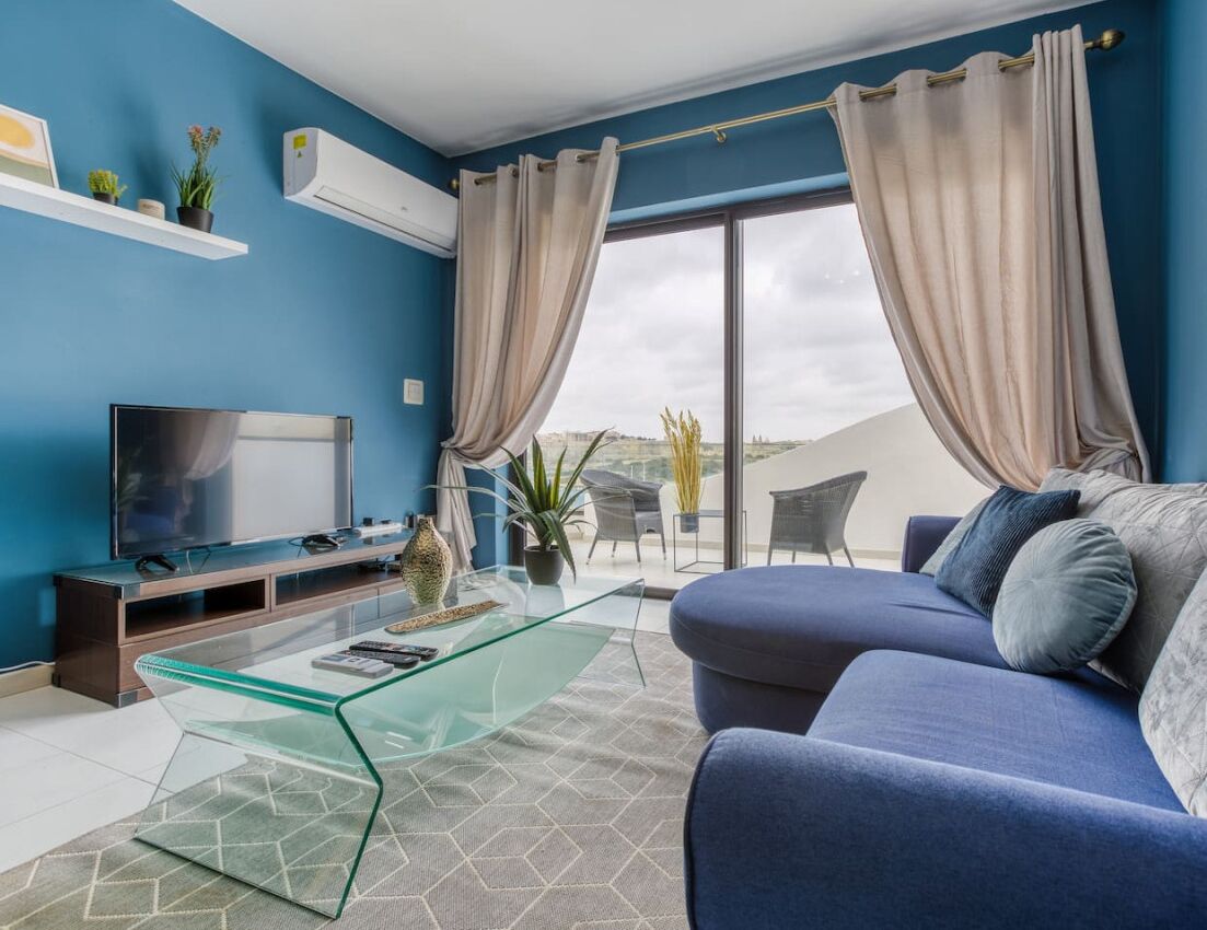 A modern living room with a blue sofa, a glass coffee table, a flat-screen TV, and a balcony with outdoor seating.
