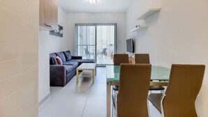 A modern living room with a sofa, coffee table, and dining area with chairs. There is a television mounted on the wall and a large window with blinds. - Cosy 1BR penthouse with Private Terrace in Gzira (Il-Gżira)
