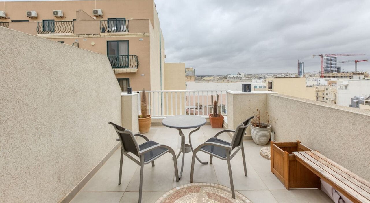 A balcony with a round table and two chairs, potted plants, and a wooden bench.