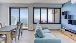 Uma sala de estar moderna com uma grande janela que oferece vista para o mar, uma área de jantar com mesa e cadeiras, e um sofá azul claro.