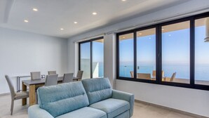 Uma sala de estar moderna com um sofá azul claro, uma mesa de jantar com cadeiras e grandes janelas com vista para o mar.